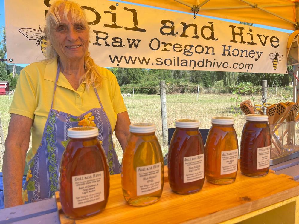 Beekeeper Sally at the Soil and Hive booth, behind a row of blue-ribbon raw Oregon honey jars under the Soil and Hive banner